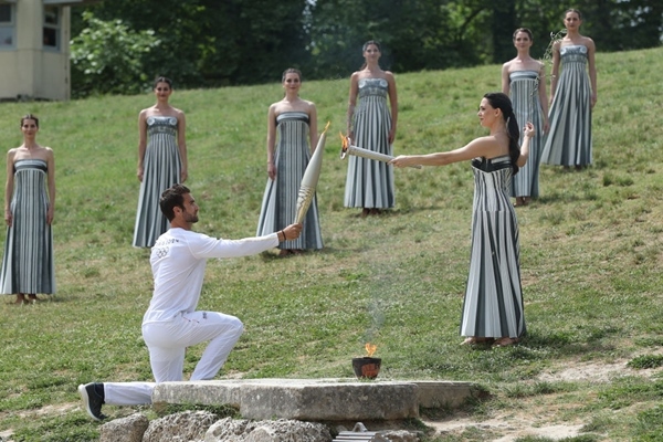 扮演最高女祭司的演員瑪麗·米娜（前右）點(diǎn)燃第一棒火炬手——希臘運(yùn)動(dòng)員、東京奧運(yùn)會(huì)男子賽艇項(xiàng)目金牌得主斯特凡諾斯·恩圖斯科斯手中的火炬。新華社記者 趙丁喆 攝